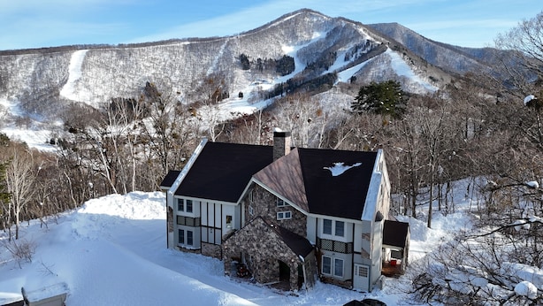 Una casa de piedra de dos plantas con techo oscuro, rodeada de terreno y árboles cubiertos de nieve, con una montaña al fondo.