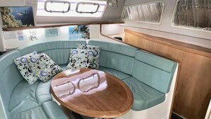 A cozy, curved seating area with teal cushions and patterned pillows, a wooden table, and a view of the ocean through a window.