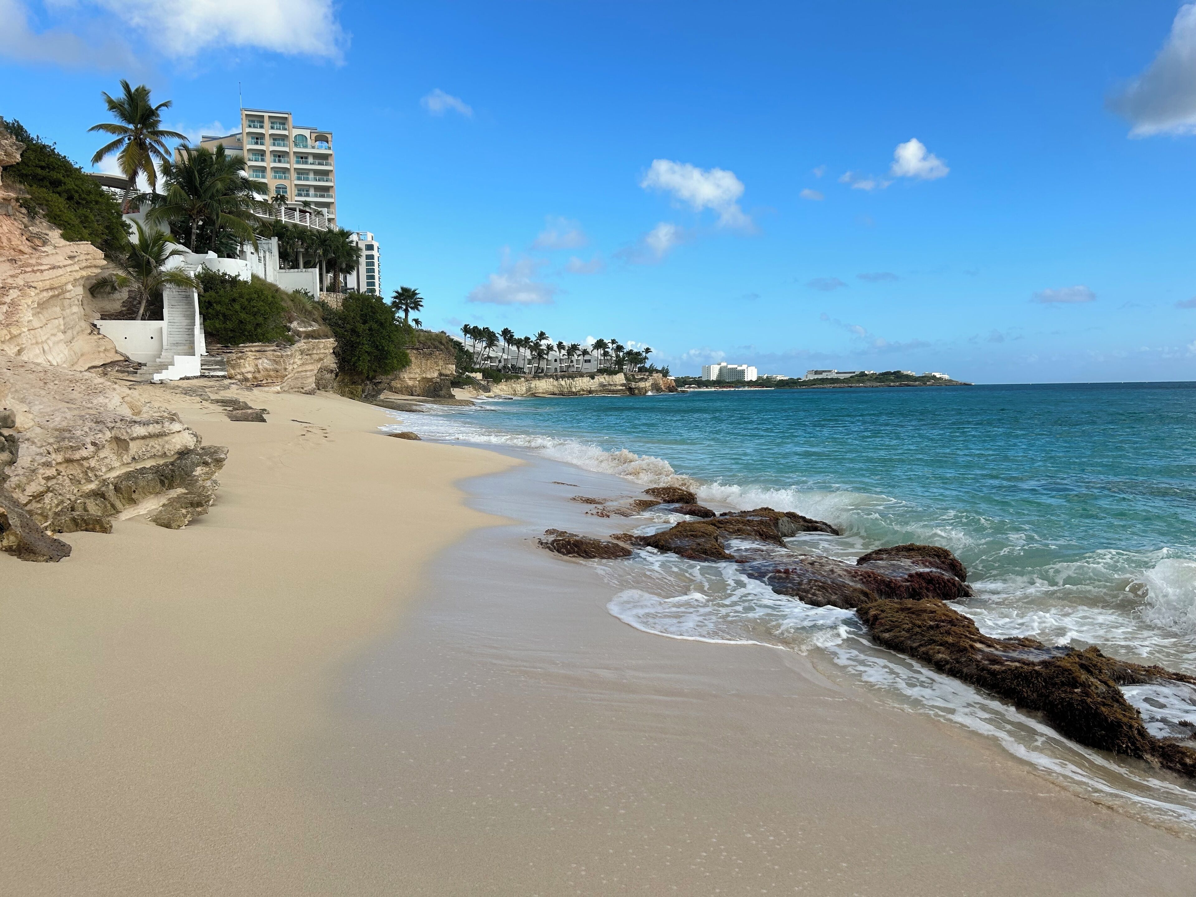 Une plage bordée d’un rivage rocheux, avec des palmiers et un bâtiment doté de balcons donnant sur la mer.