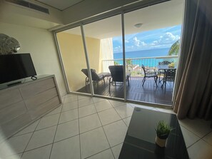 A balcony with a view of the ocean, a TV on the left, and a table with chairs.