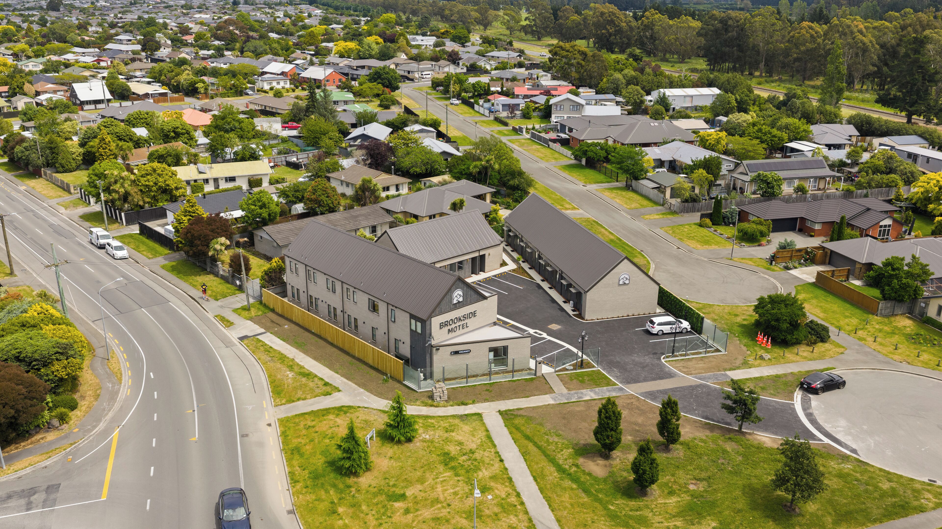 An aerial view of a residential area with a motel named "Brookside Motel" featuring a modern design, a parking lot, and a road with vehicles.
