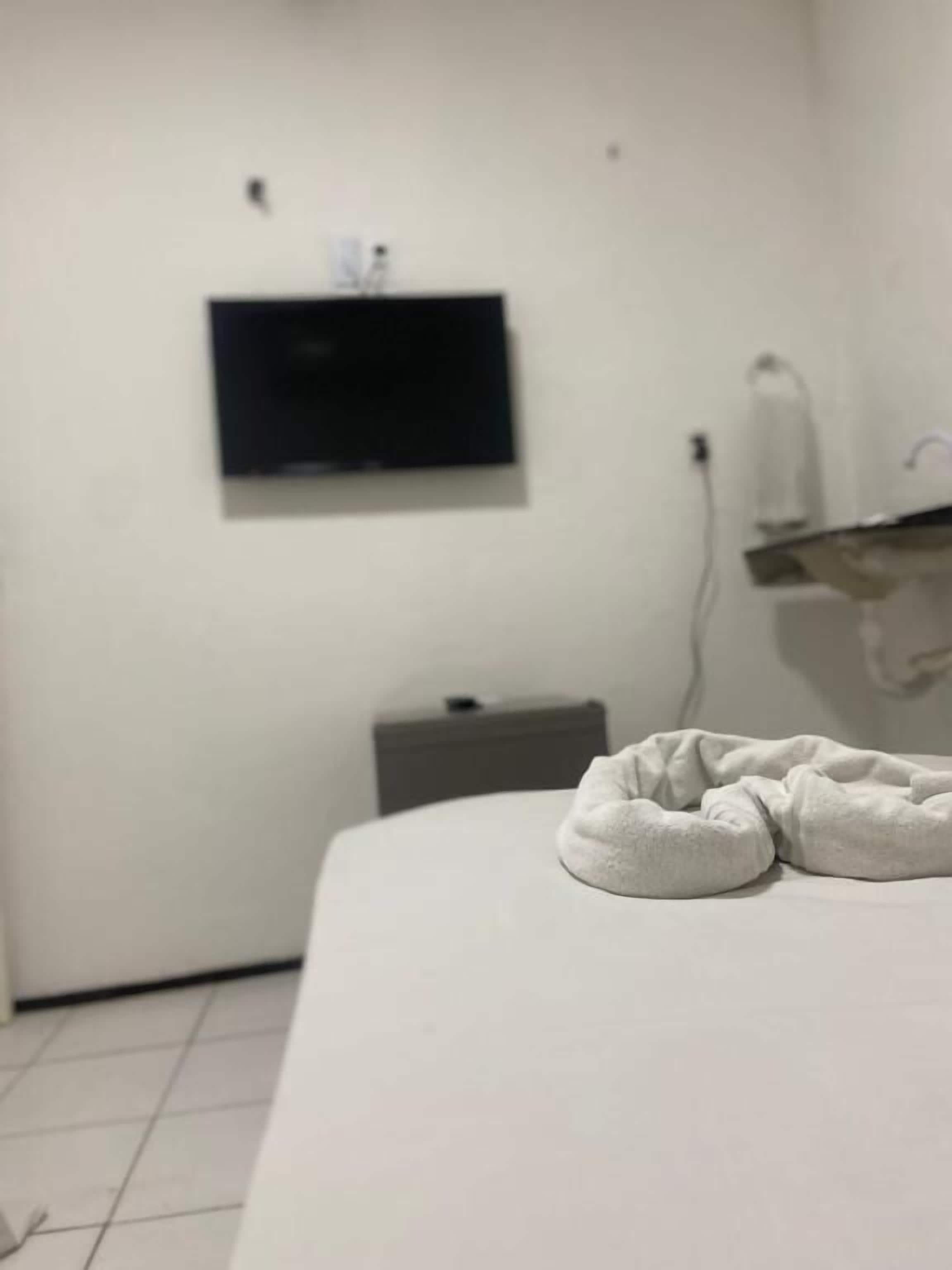 Quarto de hotel com cama, televisão presa à parede, mesa de cabeceira e toalha sobre a cama.