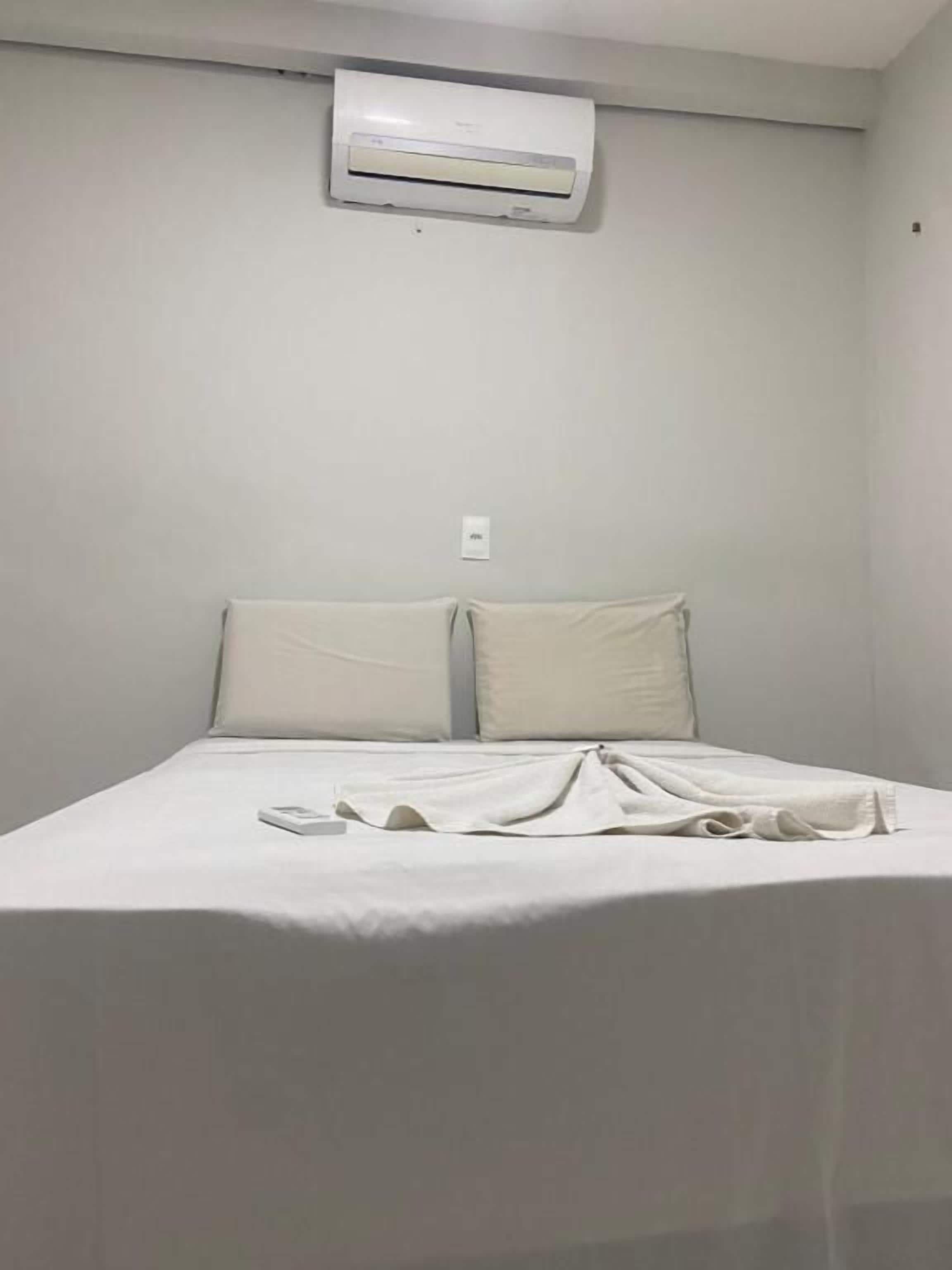 A single bed with a white mattress and pillows, an air conditioning unit mounted on the wall, and a small white switch or control panel below it.