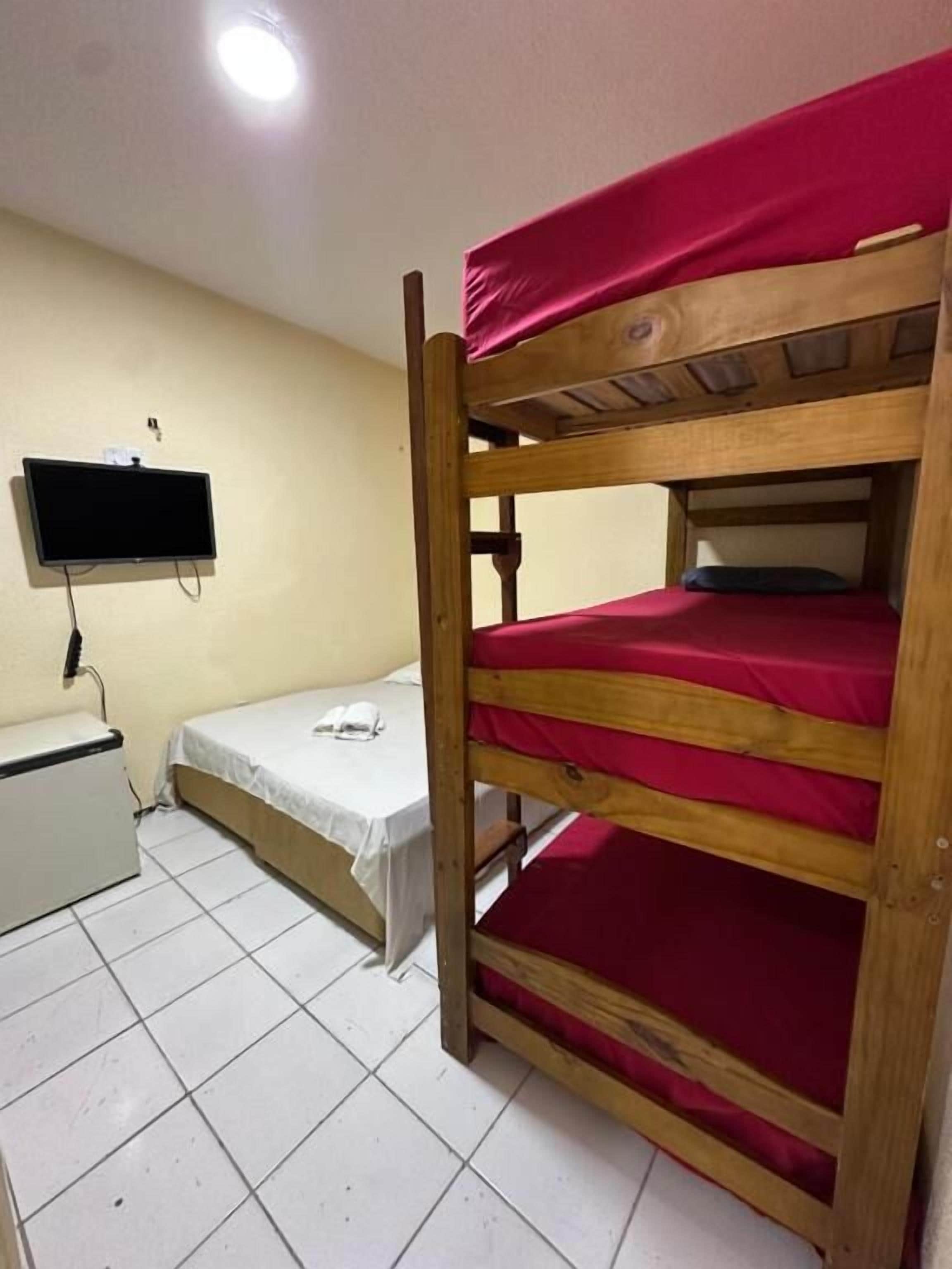 A bunk bed with red bedding, a small bedside table, a TV mounted on the wall, and a white tiled floor.