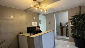 Un área de recepción moderna con recepcionista, mostrador de recepción, una planta grande en maceta y un espacio para sentarse.