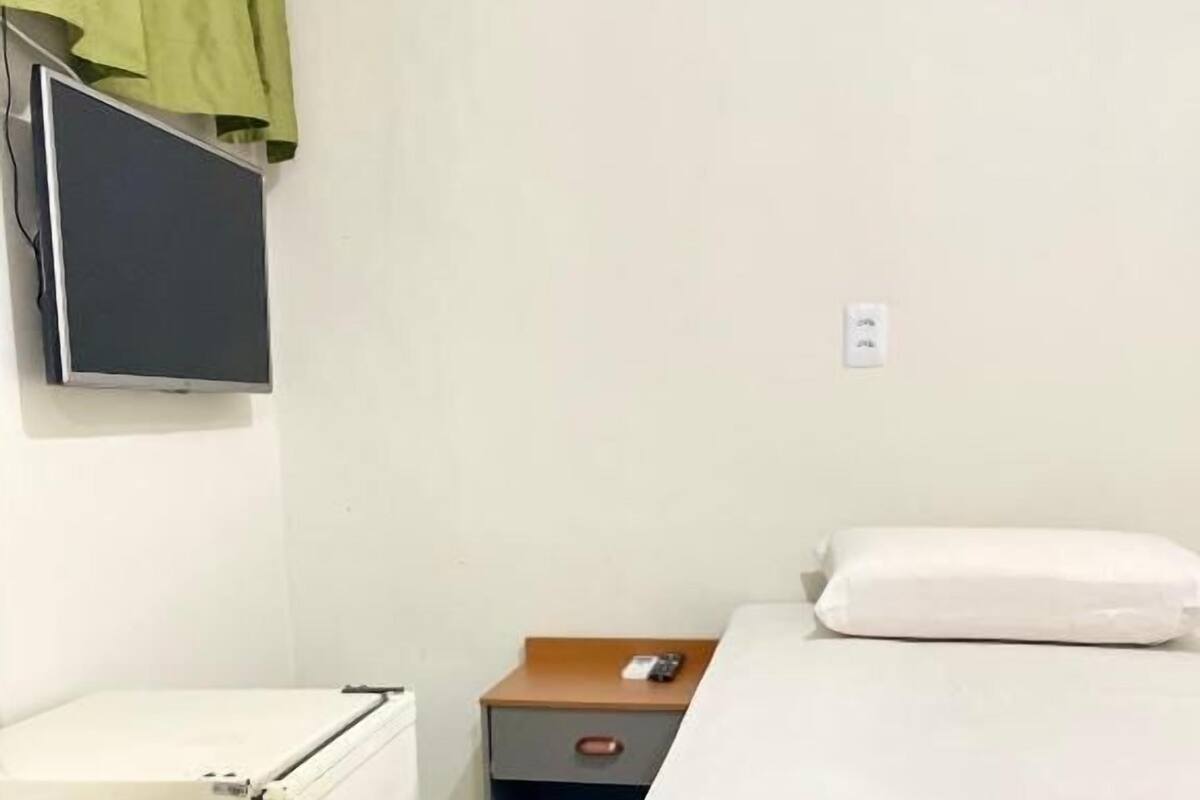 Una habitación pequeña y sencilla con una cama, una mesita de noche, una pequeña mesa y un televisor montado en la pared.