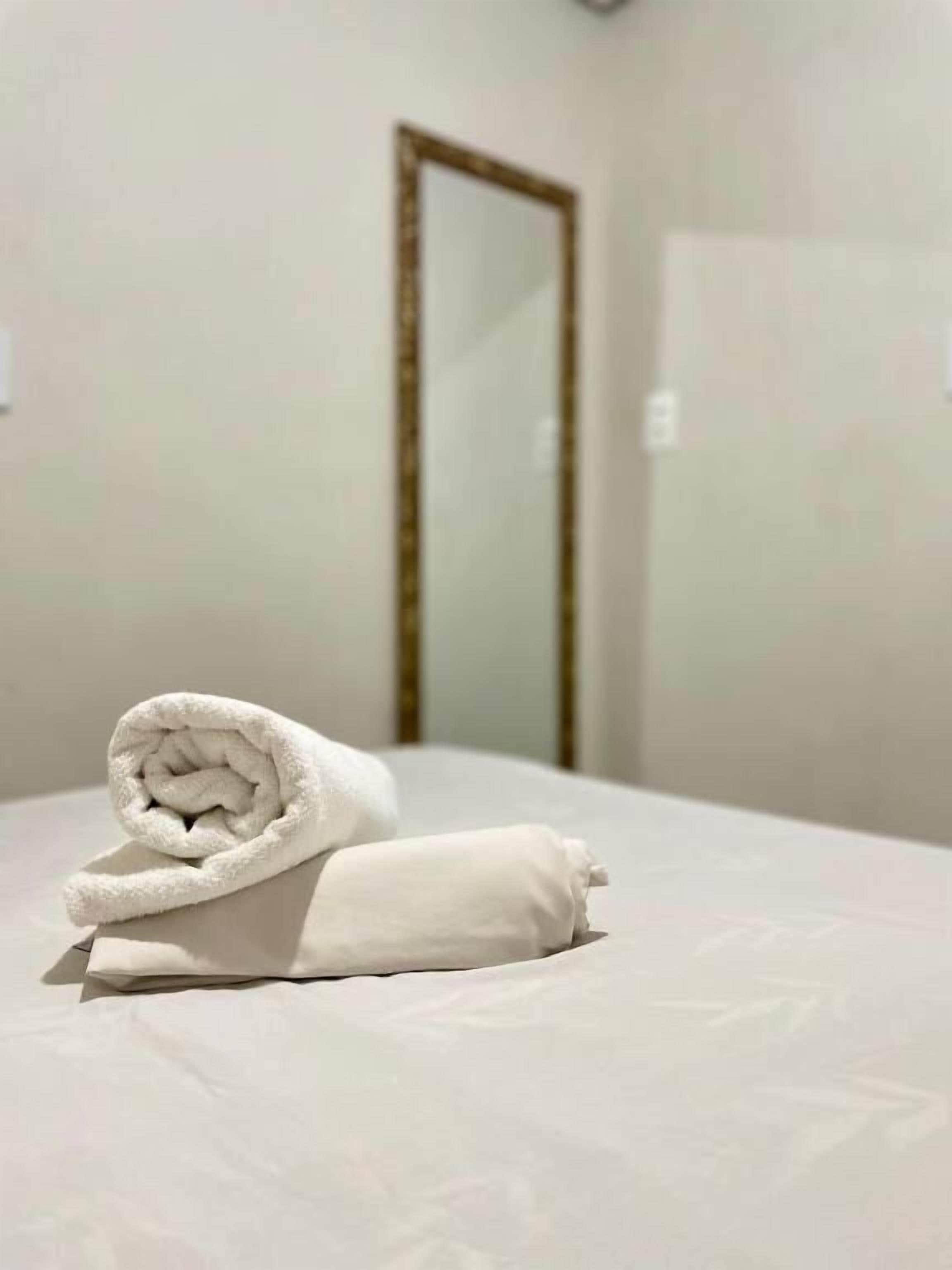 A neatly made bed with a rolled towel and a pillow in a room with a plain wall and a door in the background.