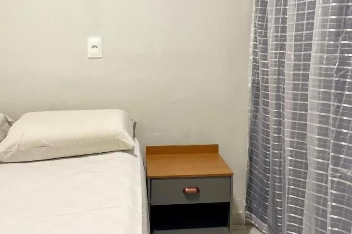 Un dormitorio con cama, una mesita de noche con cajón, una cortina y un interruptor de luz en la pared.