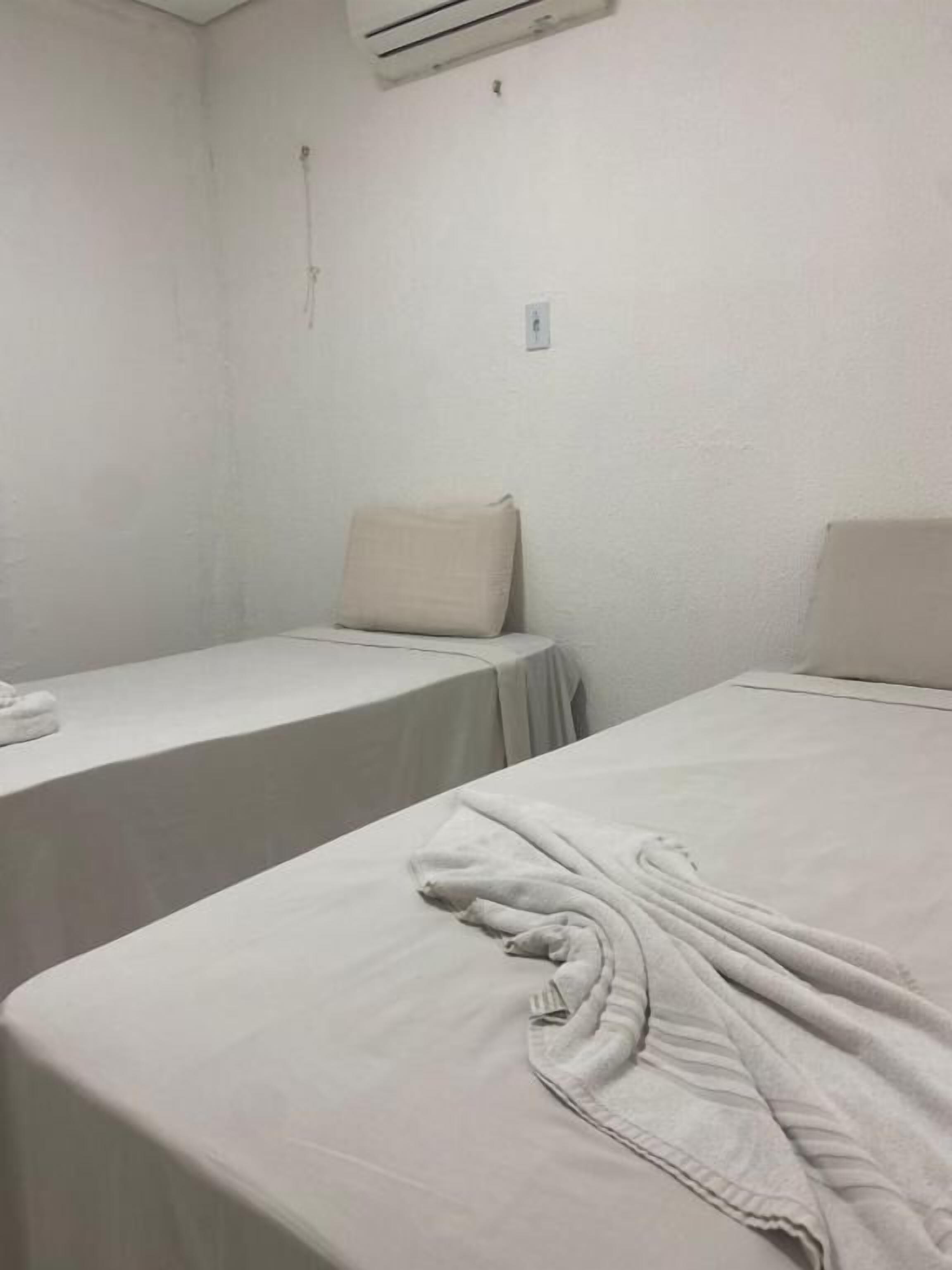 Habitación de hotel con dos camas, cada una cubierta con una colcha blanca. Una cama tiene un almohadón blanco y la otra, una bata blanca colgada. Las paredes y el techo son blancos, con un equipo de aire acondicionado montado en la pared.