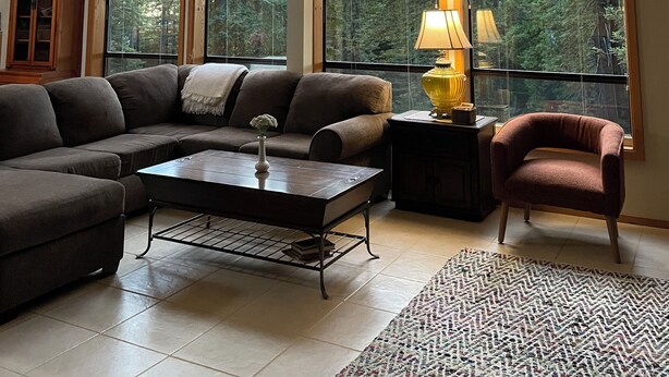 A spacious living room with a high ceiling, large windows offering a forest view, a ceiling fan, and a cozy seating area with a sofa and armchair. - Beautiful Large Home w Game Room on Private Acreage  (Gualala)