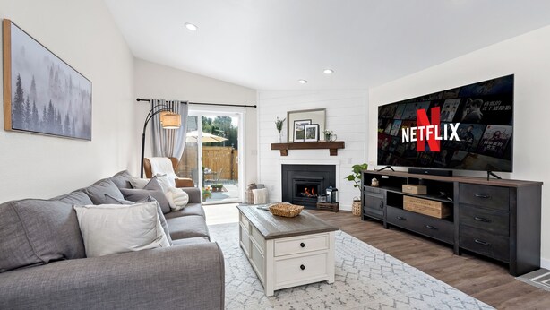 Una sala moderna con un sofá gris, una mesa de centro blanca y una chimenea. Un televisor grande está montado en la pared, y se muestra el logotipo de Netflix.