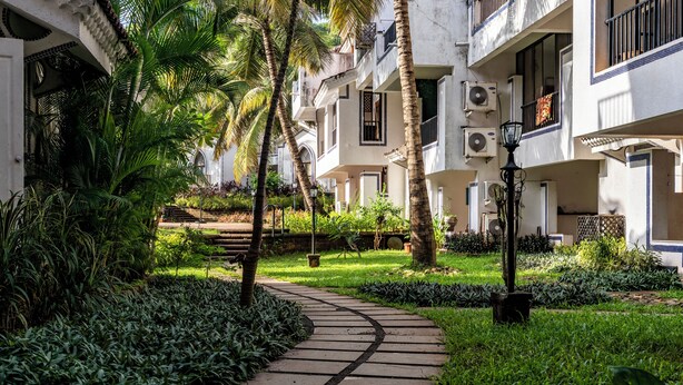 Un sentier traverse un jardin luxuriant, bordé de palmiers et de diverses plantes, et est flanqué d’immeubles à plusieurs étages dotés de balcons et de climatiseurs.