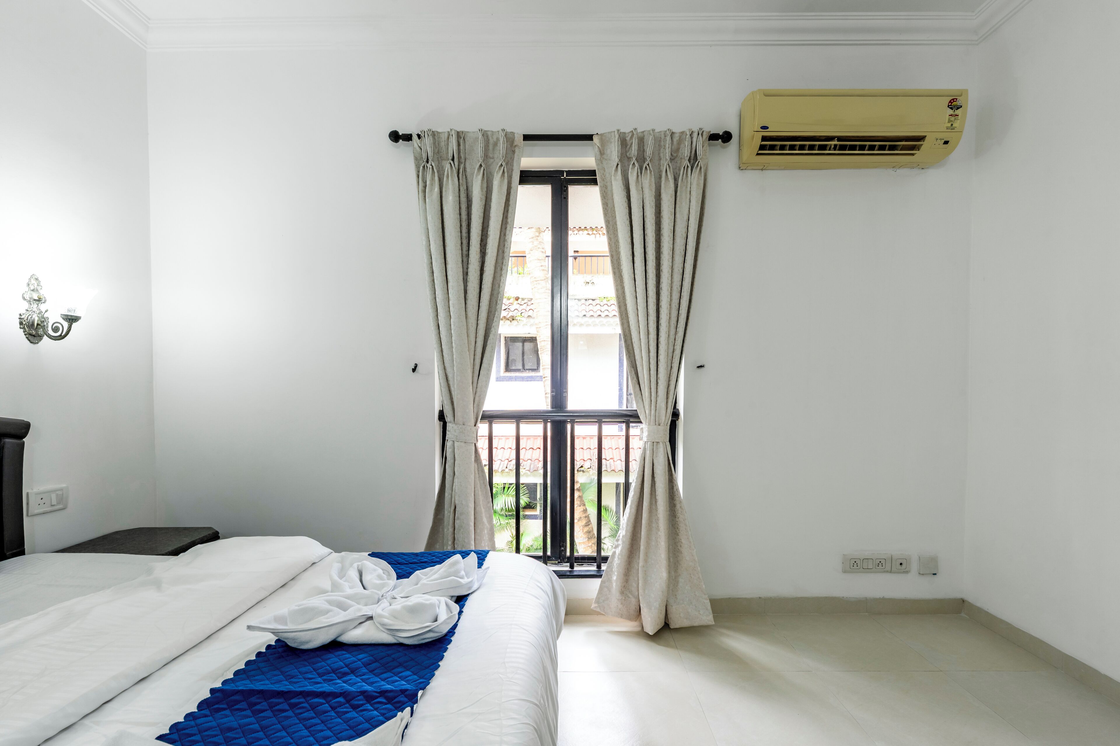 A hotel room with a bed, curtains, an air conditioning unit, and a view of the outside balcony.