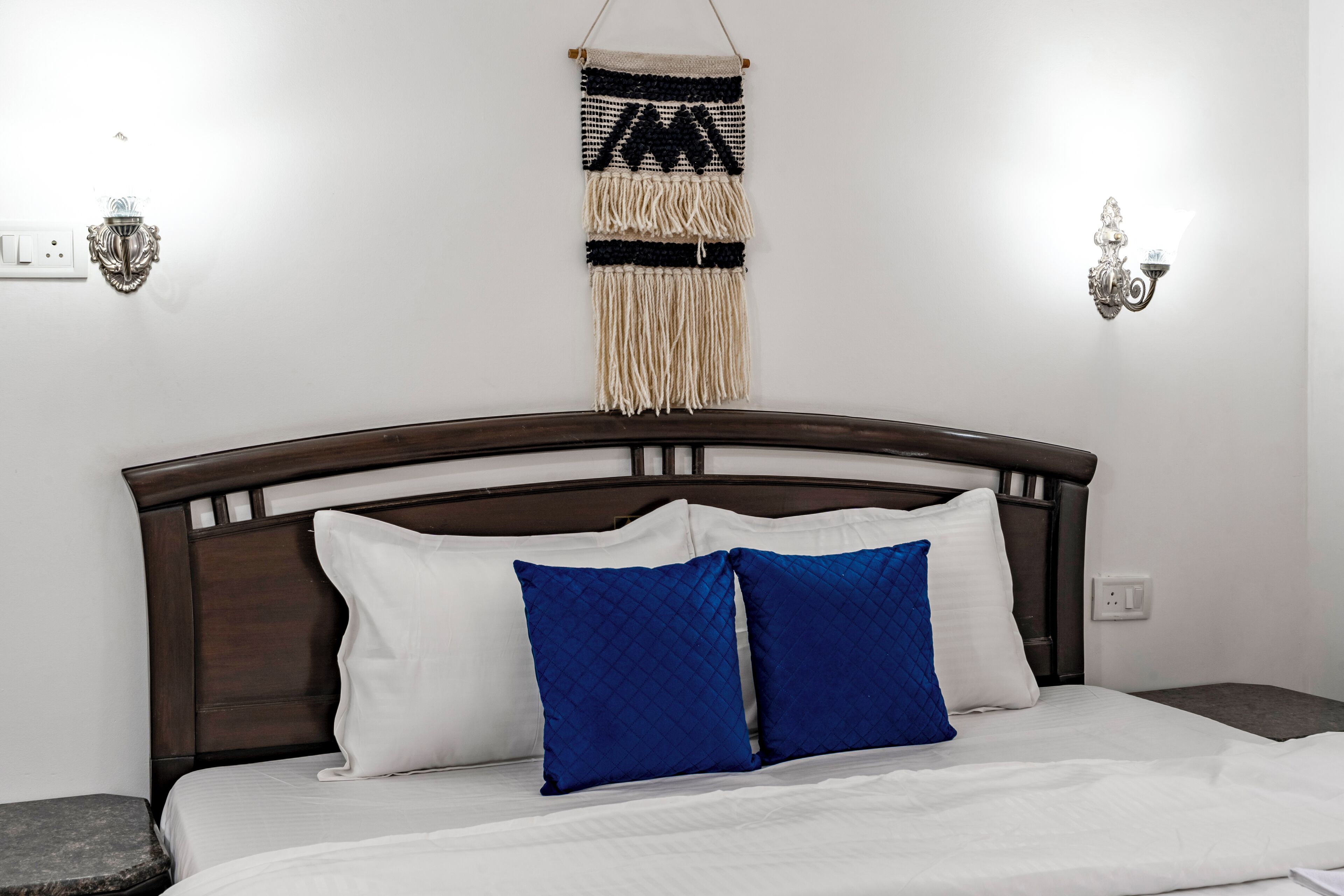 A neatly made bed with white and blue pillows, a wooden headboard, and a macrame wall hanging above.