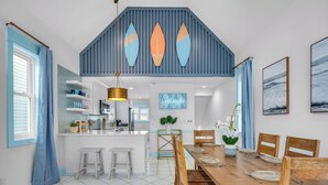 A modern kitchen with white cabinets, a central island, and a dining area with wooden chairs and a table. - Splash on St. George Island, beachfront, beautiful, close to everything (Eastpoint)