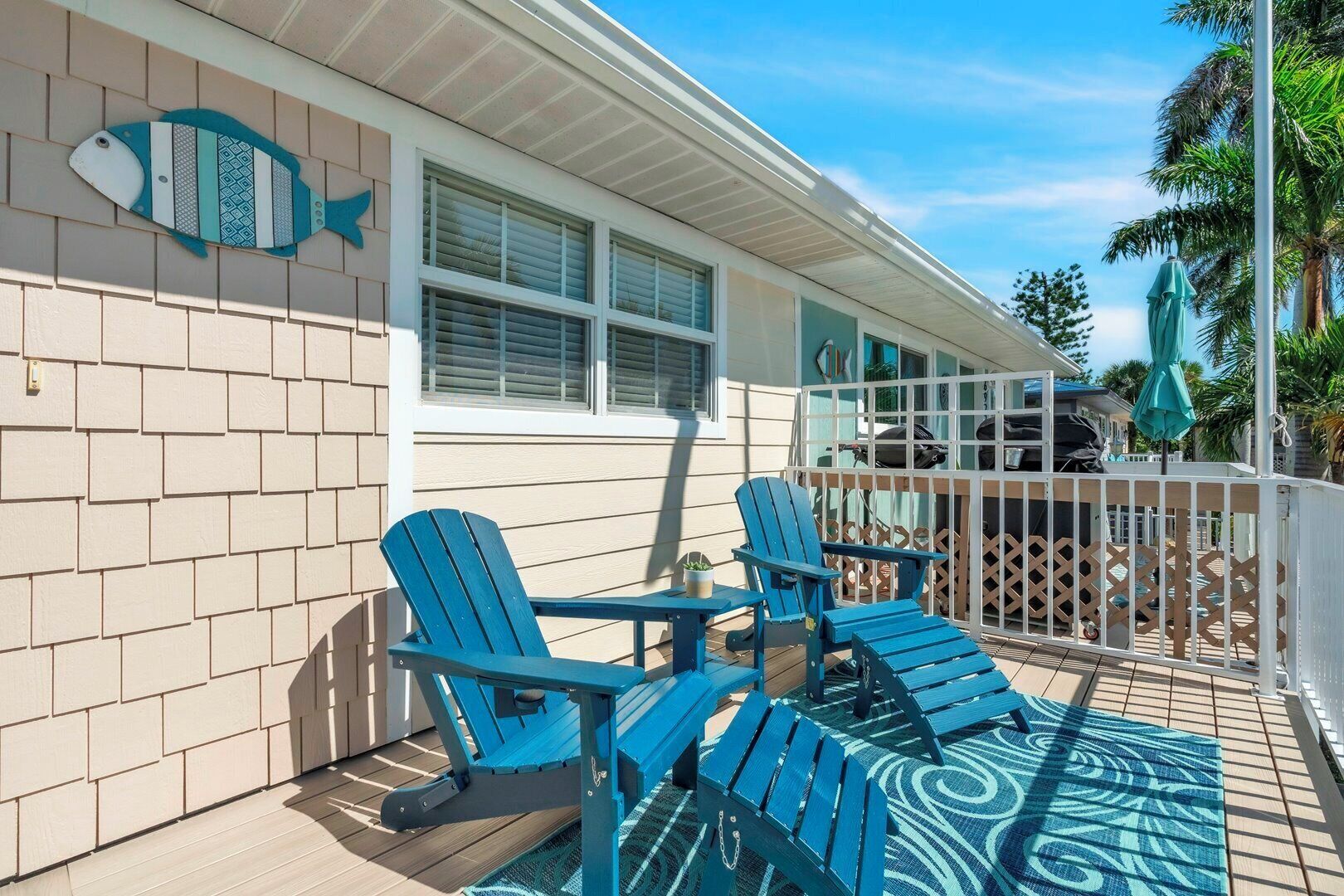 A deck with blue Adirondack chairs and a table, a white fence, and a fish-shaped decoration on the wall.