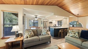 A spacious living area with a wooden ceiling, a sofa, a coffee table, and a dining table. A kitchen with a bar and a refrigerator is visible in the background. - Fairway Village 25 - SHARC + Condo Pool, Hot Tub, Near Golf Course, A/C (Sunriver)