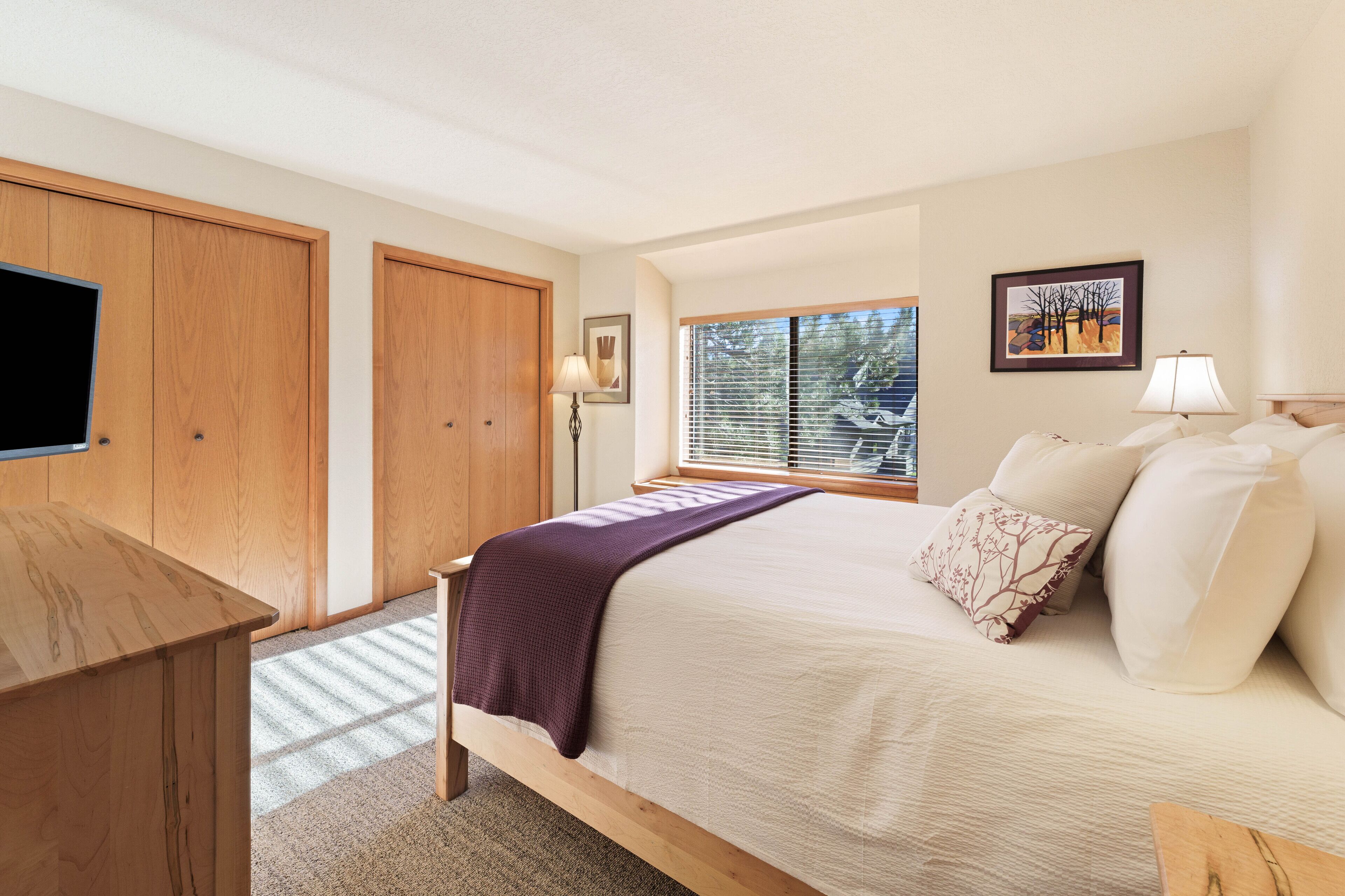 Une chambre à coucher comprenant un lit, une armoire en bois, un bureau, une fenêtre avec des stores et un téléviseur fixé au mur.