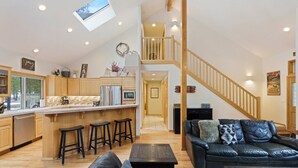 A spacious living area with a high ceiling, a kitchen with wooden cabinets, a dining area with stools, and a staircase leading to an upper level. - Cultus 15 - Beautiful, SHARC, Pool Table, A/C, Gas Fireplace (Sunriver)