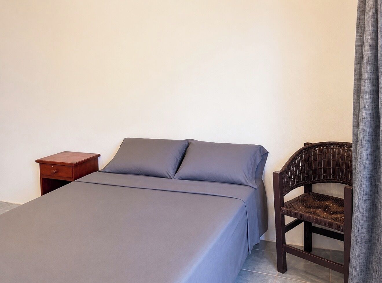 Una cama individual con colcha gris, una mesita de noche de madera con cajón y una silla de mimbre.