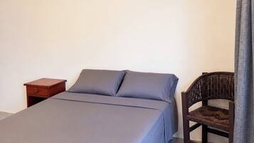 Una cama individual con colcha gris, una mesita de noche de madera con cajón y una silla de mimbre.