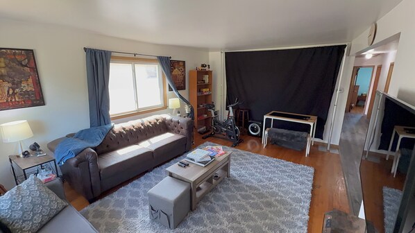 A living room with a sofa, a coffee table, a lamp, a bookshelf, and a large window with curtains. - Quiet Retreat in Morgan Heights, Sleeps 6, 1.5 baths (Milwaukee)