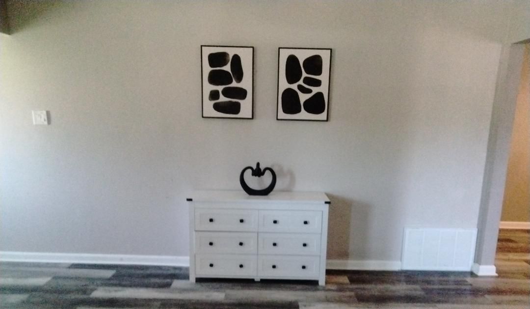 Une pièce avec une commode blanche, deux œuvres d’art abstraites accrochées au mur et un tapis à motifs.