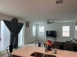 A modern living room with a large TV, a ceiling fan, and a dining area with a sink.