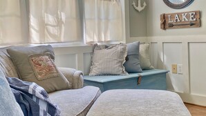 A cozy room with a blue sofa, a window with curtains, a wall-mounted anchor decoration, and a 'LAKE' sign.
