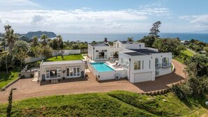 A modern, multi-story house with a large swimming pool, surrounded by lush greenery and a view of the ocean.