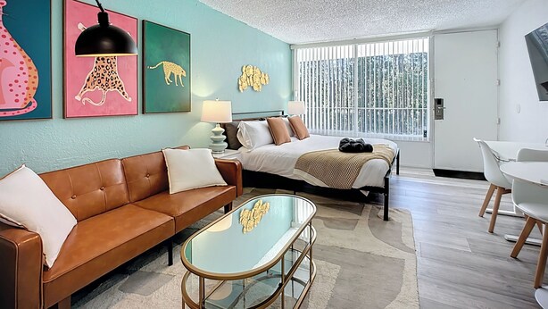 A modern hotel room with a brown leather sofa, a glass coffee table, a bed with a white and brown striped blanket, and a large window with blinds. - Cozy King Studio | 5 Min to Zoo, JU & DT (Jacksonville)