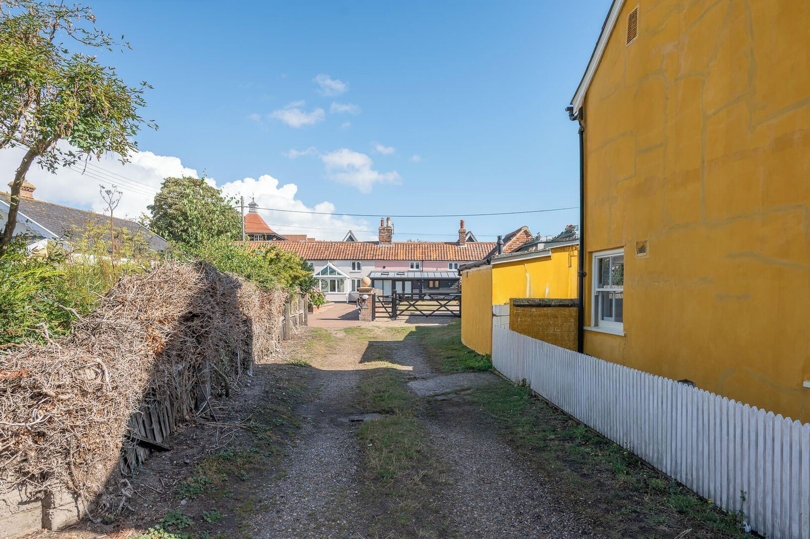 A narrow gravel path leading to a yellow building with a white fence, surrounded by vegetation and a distant house with a red roof.