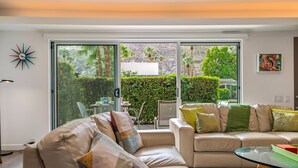 Un salon moderne avec un canapé beige, une table basse en verre et une porte-fenêtre coulissante donnant sur une terrasse agrémentée de verdure et d’une structure blanche.