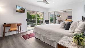 Une chambre dotée d’un grand lit, d’un bureau et d’une chaise, d’un ventilateur de plafond, et offrant une vue sur les palmiers et les montagnes grâce à de grandes fenêtres.