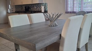A modern dining area with a large wooden table, white upholstered chairs, and a kitchen in the background with a geometric backsplash.
