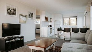 Un salon moderne avec un téléviseur à écran plat, une table basse en bois et un canapé gris. Il y a également un coin cuisine équipé d’un réfrigérateur, d’une cuisinière et de tabourets de bar.
