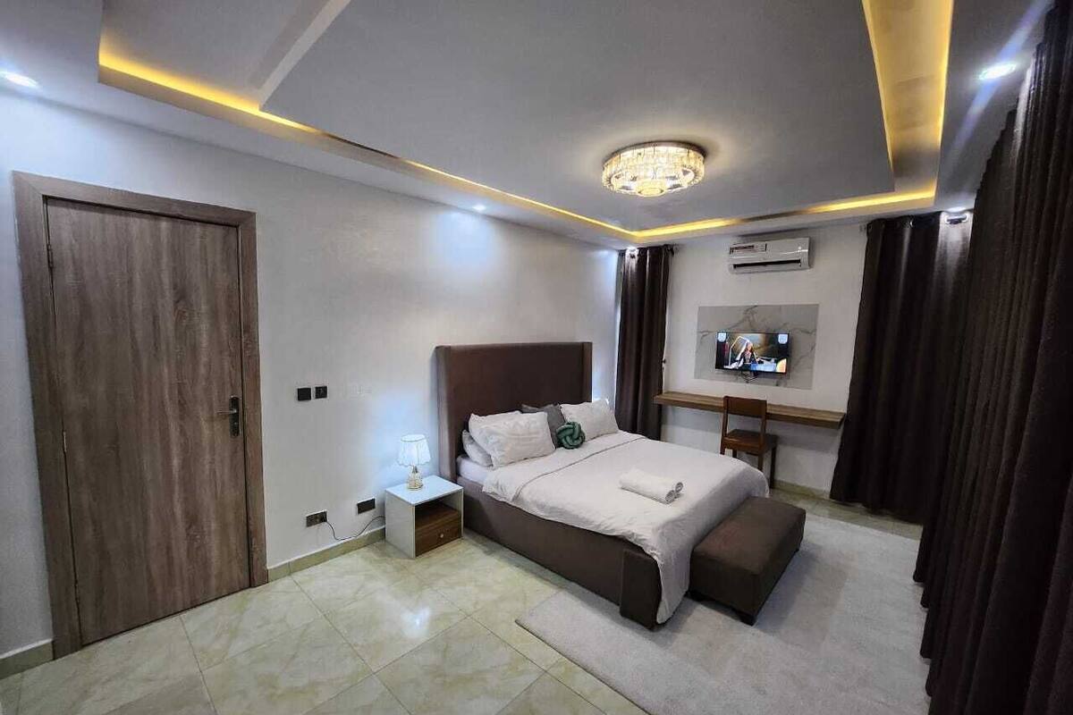 A modern hotel room with a large bed, a bedside table, a TV, and a desk with a chair.