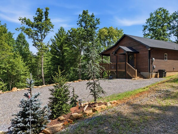 A log cabin with a porch, surrounded by trees and a gravel driveway.
