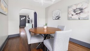 Un coin repas comprenant une table ronde en bois, quatre chaises grises à revêtement rembourré, un lustre blanc et deux tableaux représentant des motifs floraux accrochés au mur.