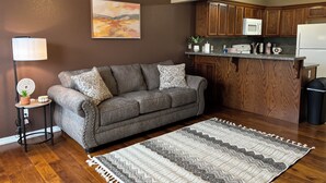 A living room with a gray sofa, patterned cushions, a wooden floor, a kitchen with wooden cabinets, a microwave, and a coffee maker. - New 2 bedroom home near Downtown (West Plains)