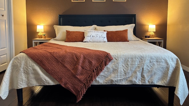 A neatly made bed with a brown blanket, white pillows, and a decorative pillow with text. There are two bedside lamps, a wall clock, and three framed pictures above the bed. - New 2 bedroom home near Downtown (West Plains)