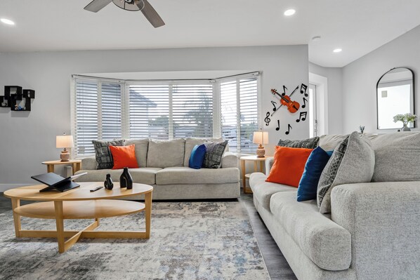 A modern living room with a sofa, coffee table, and lamps. There are windows with blinds, a ceiling fan, and a wall with a musical note decoration. - Luxe by Jenn • 3BR Retreat w/ Heated Pool (Lauderdale Lakes)