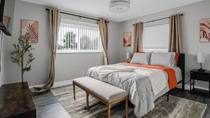 Un dormitorio con cama, banco, mesita de noche, lámpara y planta en maceta.