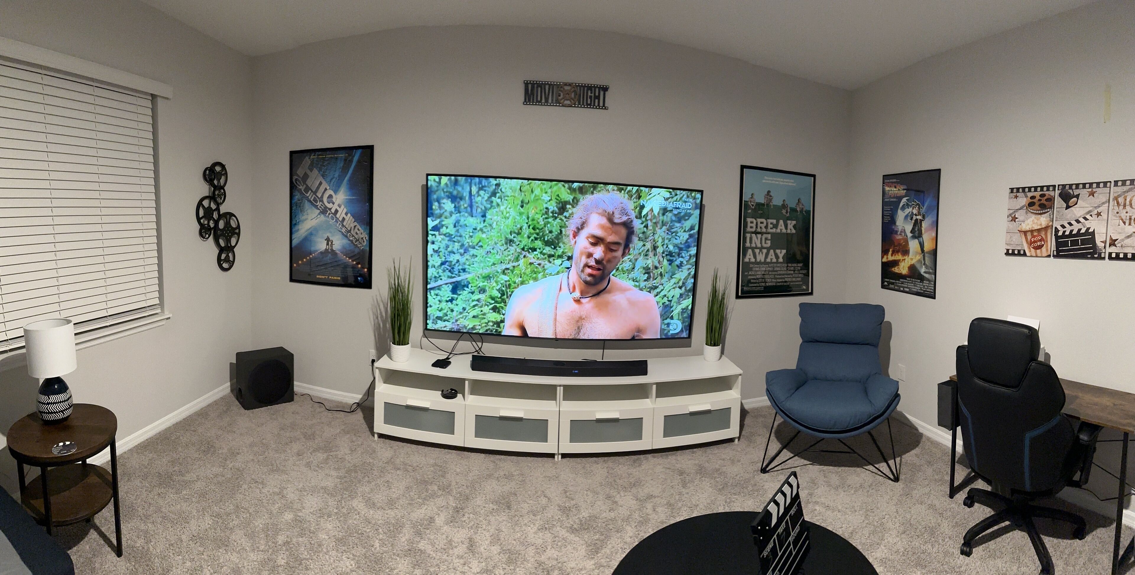 Una sala de estar con un televisor de pantalla plana grande, un mueble blanco para entretenimiento, dos sillas azules, una mesita con una lámpara y carteles de películas enmarcados en la pared.