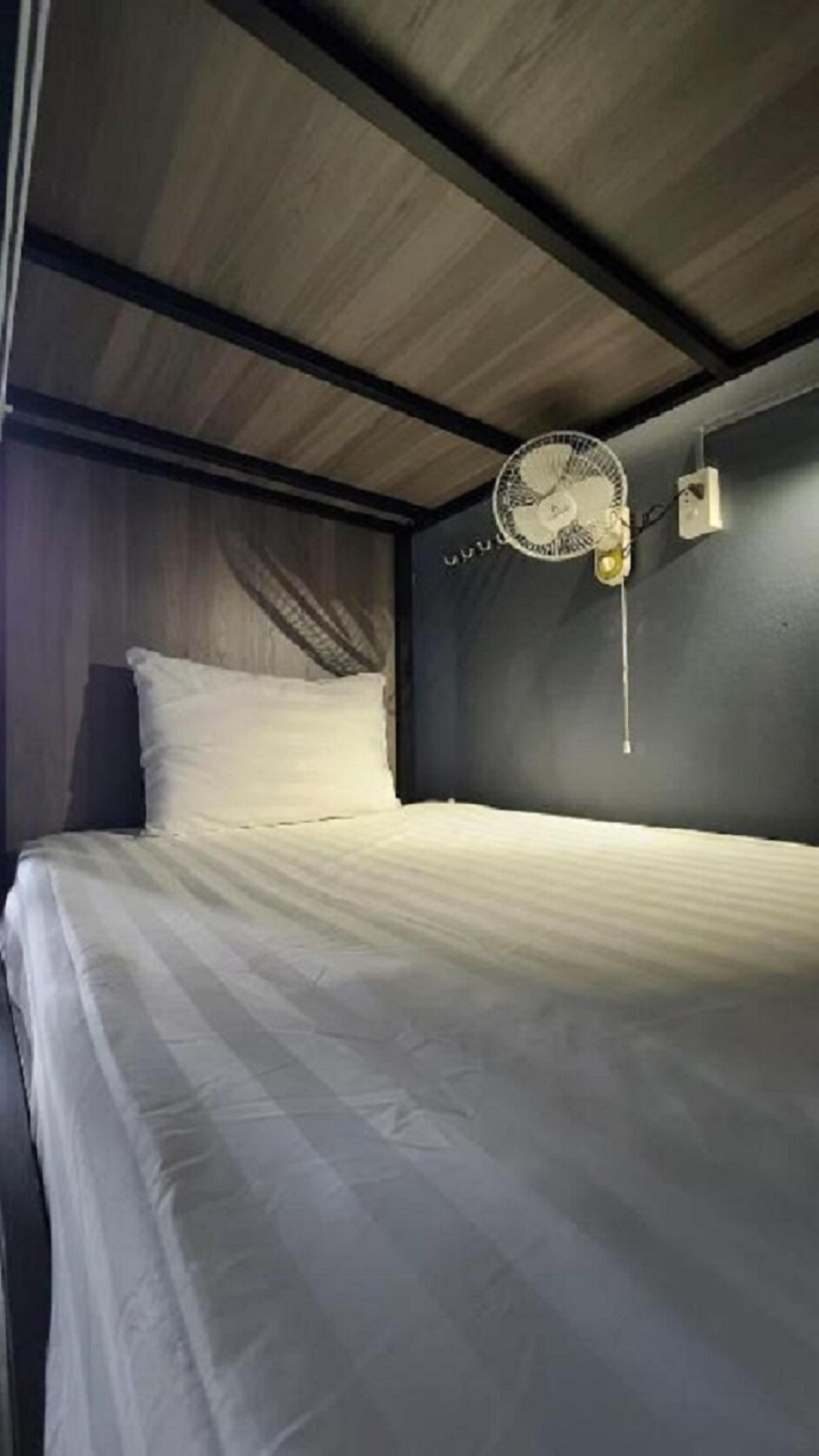 A single bed in a room with a dark ceiling and walls, a wall-mounted fan, and a small light fixture.