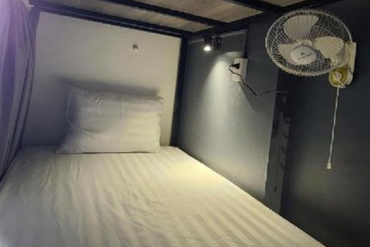 Quarto pequeno e com pouca luz, com uma cama de solteiro, um ventilador na parede e uma luminária.