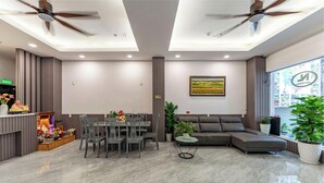 A modern hotel lobby with a dining area, a sofa, ceiling fans, and a large window with a view.