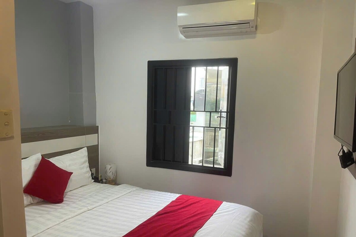 A single bed with white bedding and a red accent, a window with black bars, and a wall-mounted air conditioner.