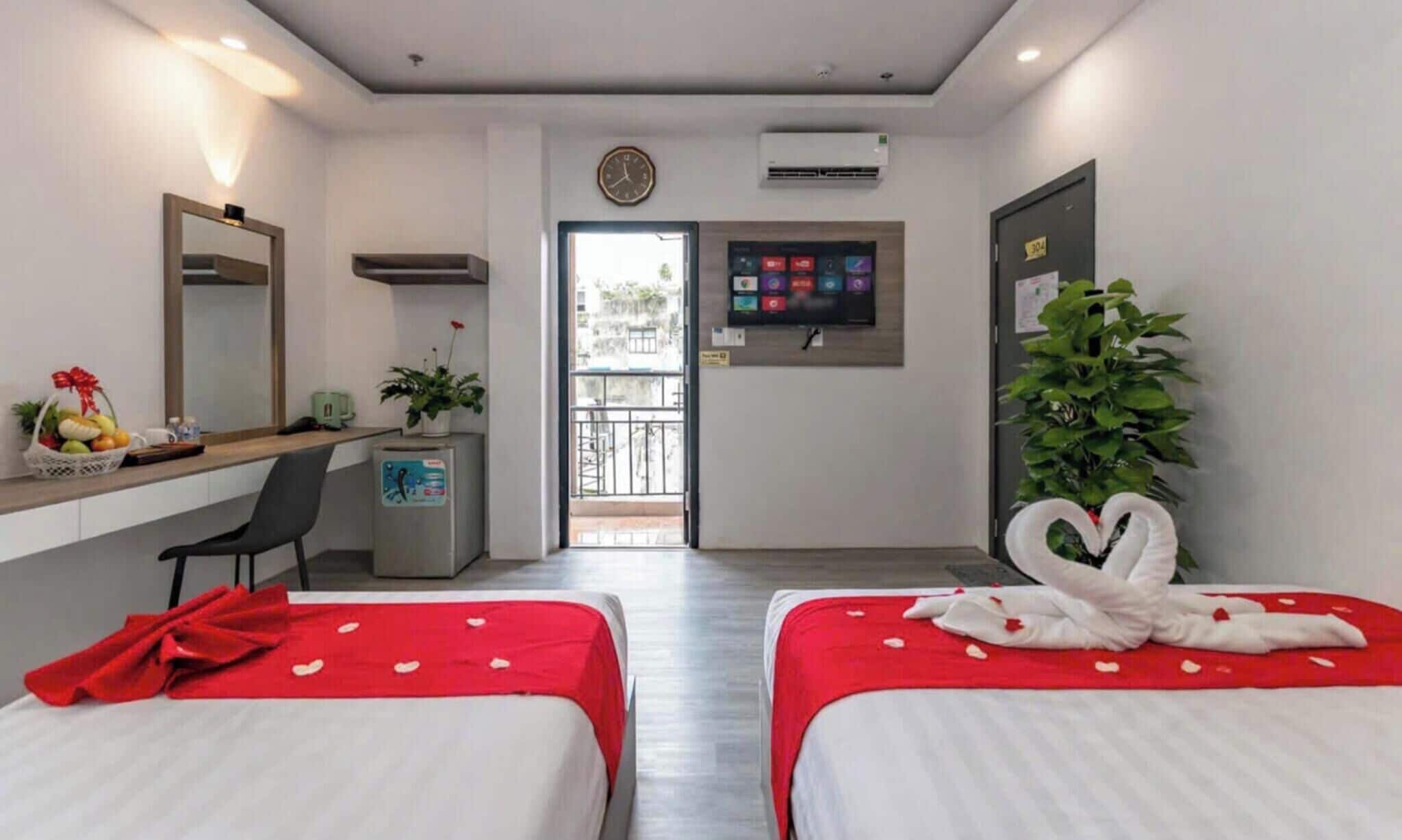 Habitación de hotel con dos camas, un escritorio con computadora, un refrigerador pequeño, un reloj y un equipo de aire acondicionado, además de una puerta con número.