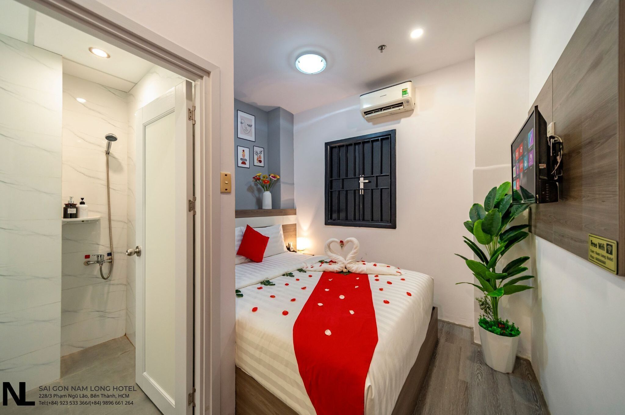 Et hotelværelse med brusebad, en seng med rødt og hvidt sengetæppe, en potteplante og en aircondition-enhed på væggen.
